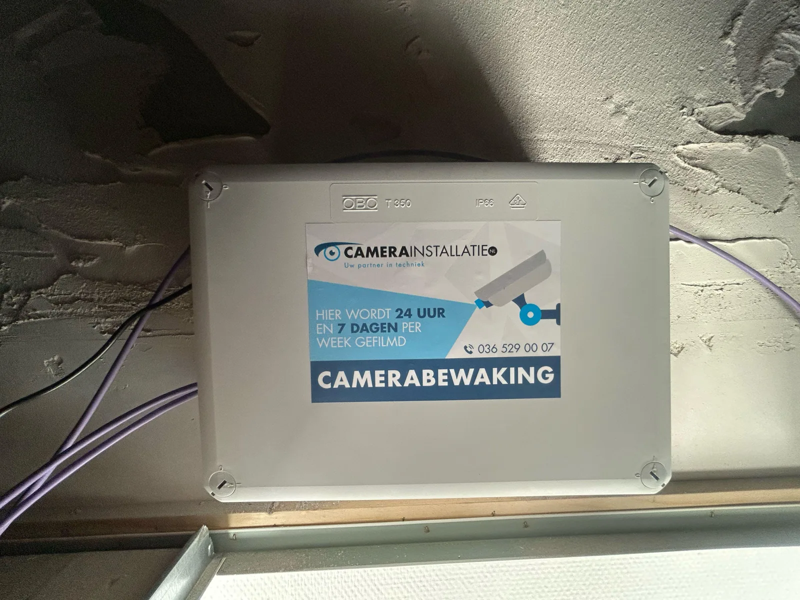 Grote grijze lasdoos boven het plafond met een sticker van CameraInstallatie.nl, waarin de UniFi Door Hub zit, met de paarse netwerkkabels ernaast