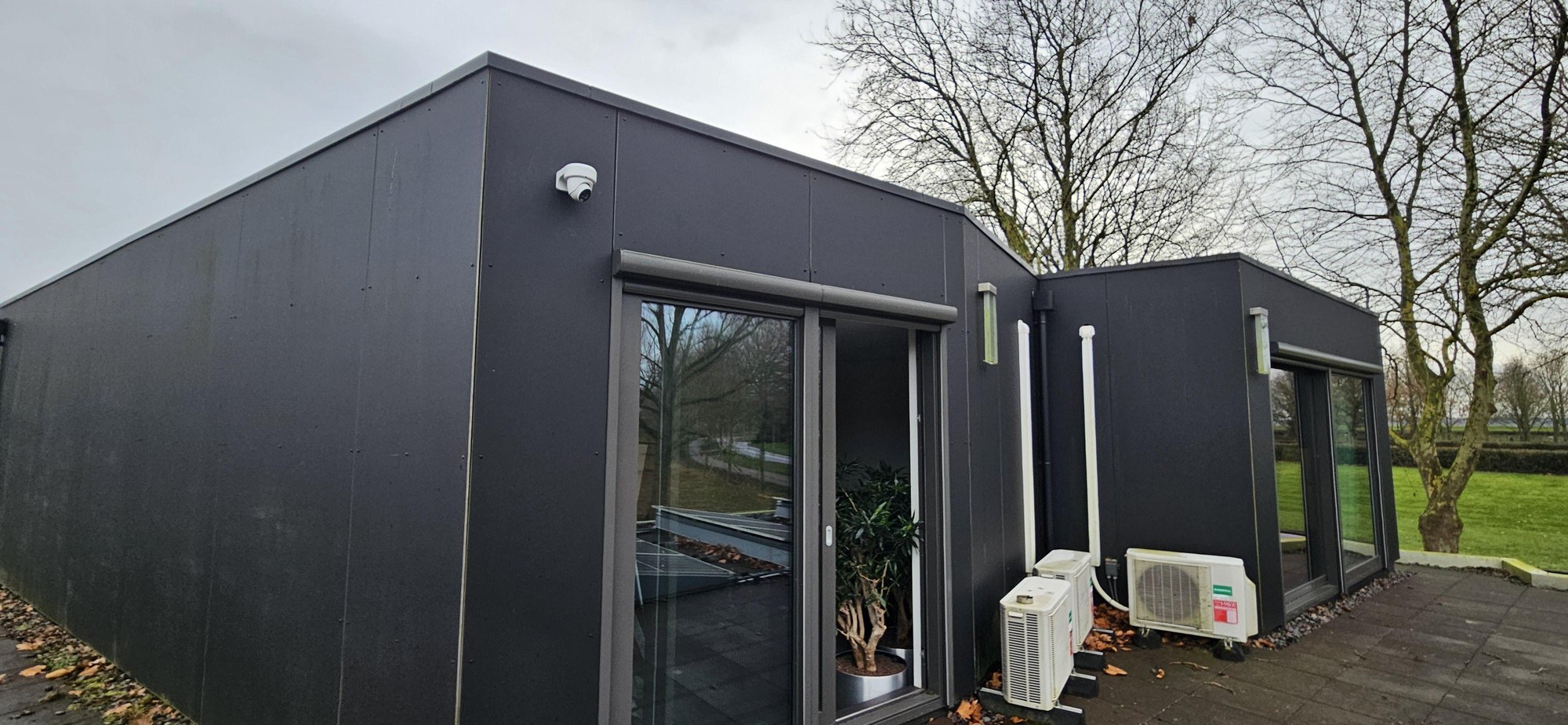 Moderne woning met donkere gevelbekleding en grote ramen, met een witte camera onder de dakrand