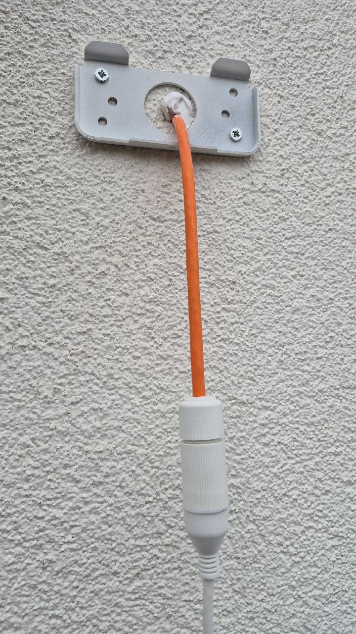 Lege muurbeugel op een witte gevel met een oranje kabel door het midden en een afgesloten aansluiting eronder