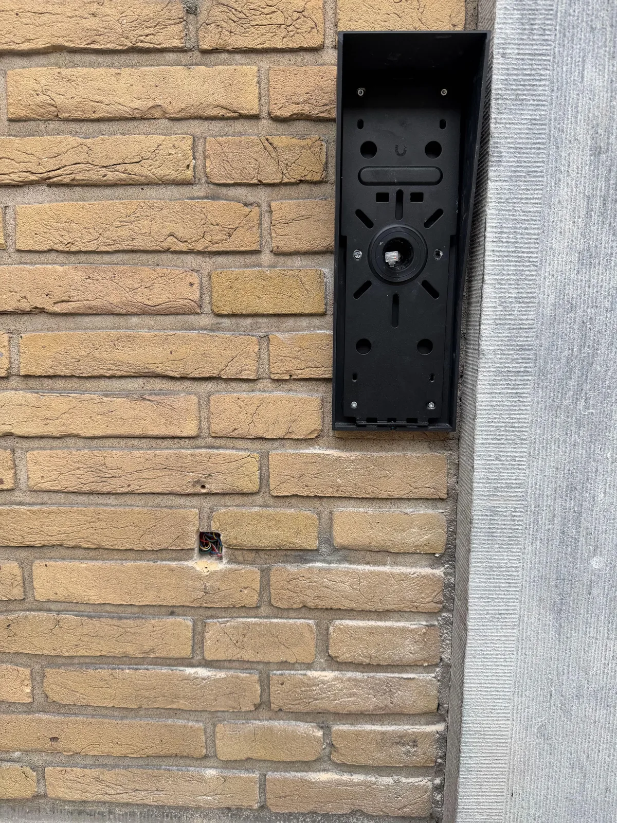 Zwarte opbouwbak met de montageplaat van de UniFi-intercom op een gele bakstenen gevel
