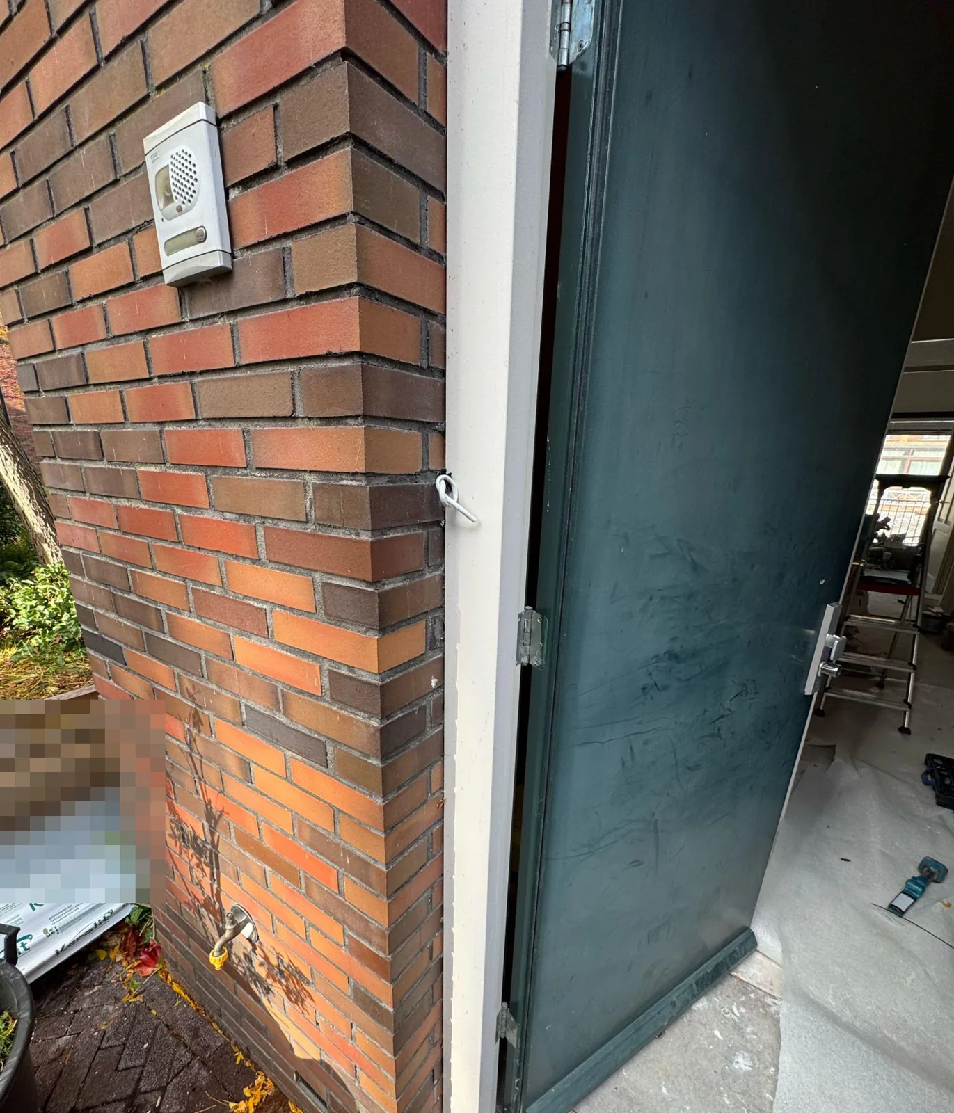Openstaande voordeur tijdens de verbouwing, met beschermfolie op de vloer en materiaal binnen, naast een bakstenen gevel