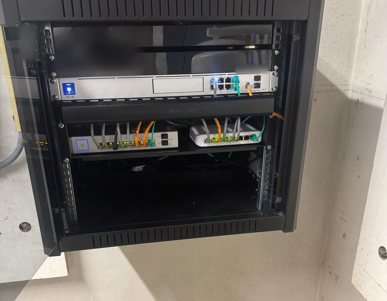 Open zwarte patchkast van 9U aan een betonwand met een UniFi Dream Machine SE en twee switches met patchkabels
