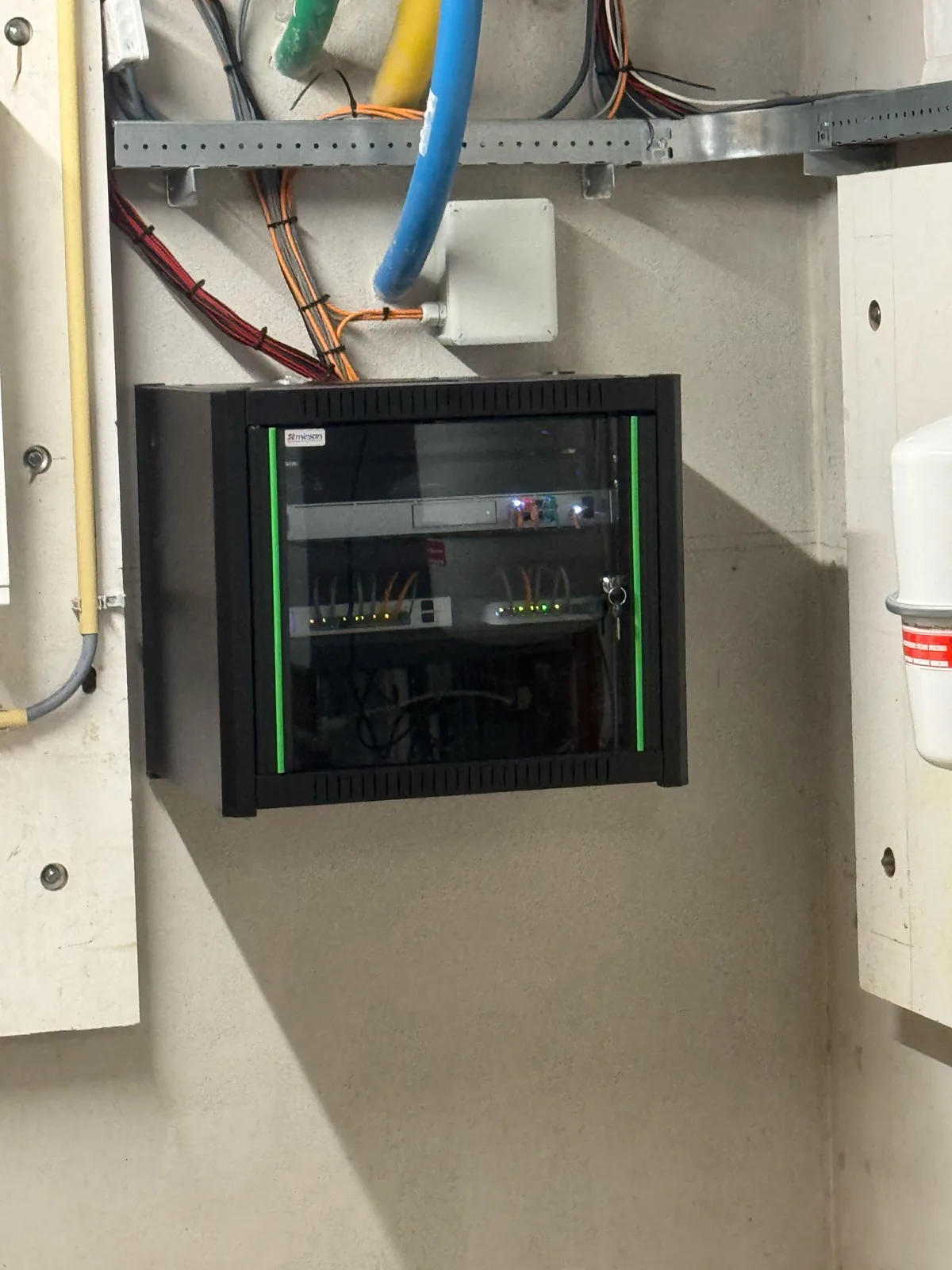 Gesloten patchkast met glazen deur aan de wand van een technische ruimte, met kabelgoot en invoer erboven