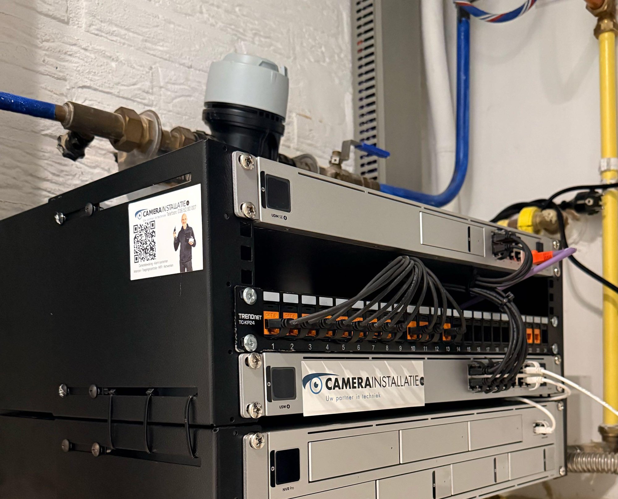 Zwarte wandkast met recorder, patchpaneel met gelabelde kabels en switch, met de sticker van CameraInstallatie.nl