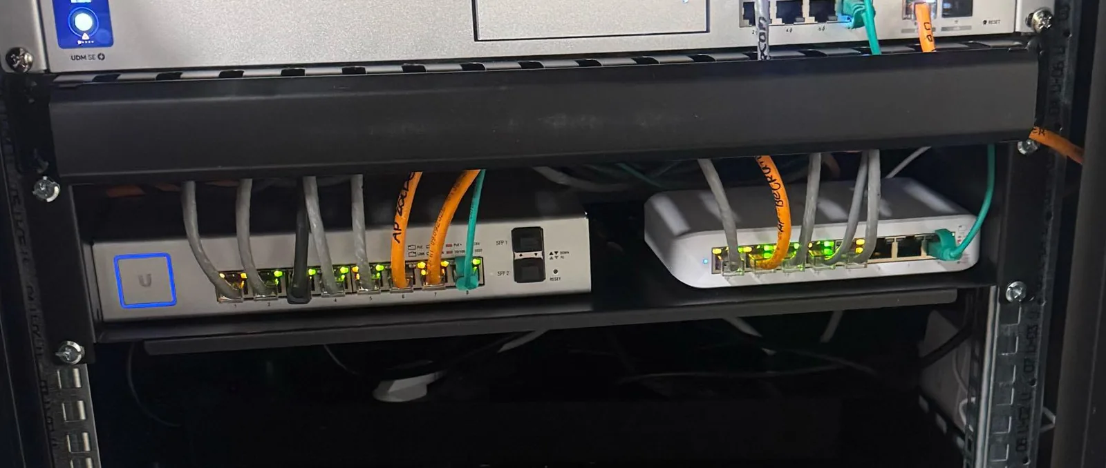 Twee UniFi-switches op een legbord in de patchkast met korte grijze en oranje patchkabels