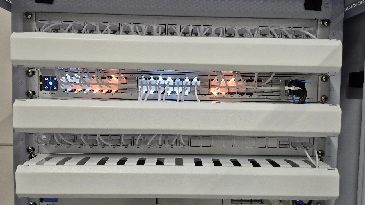 UniFi-switch met verlichte poorten tussen twee patchpanelen en kabelmanagement in de patchkast