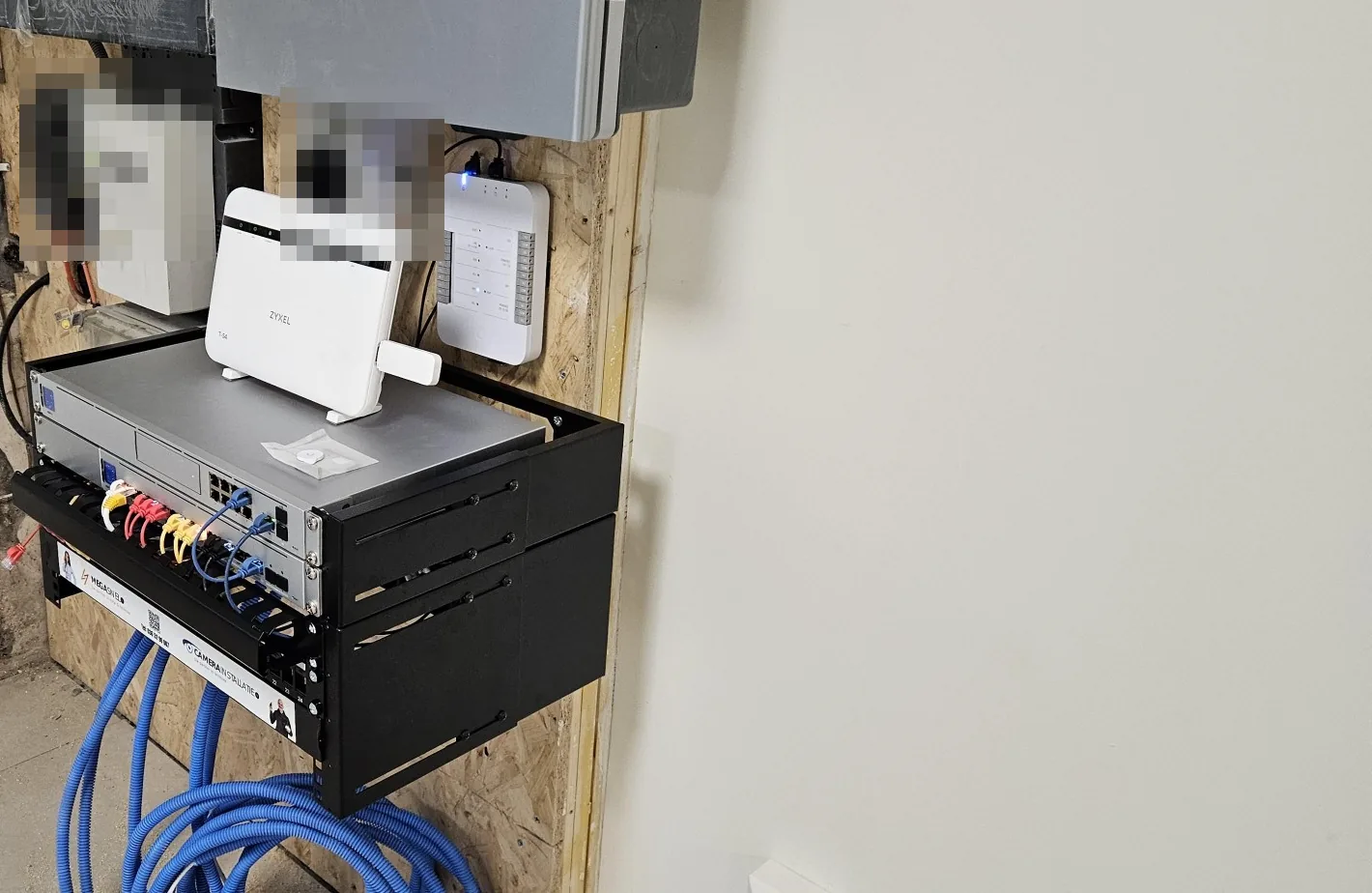 Patchrek in de kelder van opzij gezien, met router, switch, patchpaneel en blauwe kabels die van onderen binnenkomen