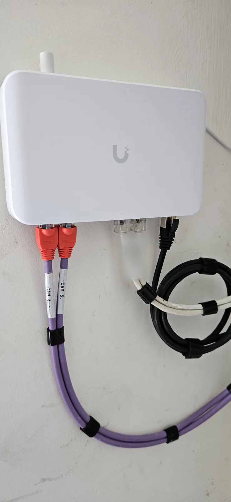 Witte UniFi PoE-switch aan de wand, met paarse netwerkkabels met labels CAM 4 en CAM 5, gebundeld met zwart klittenband