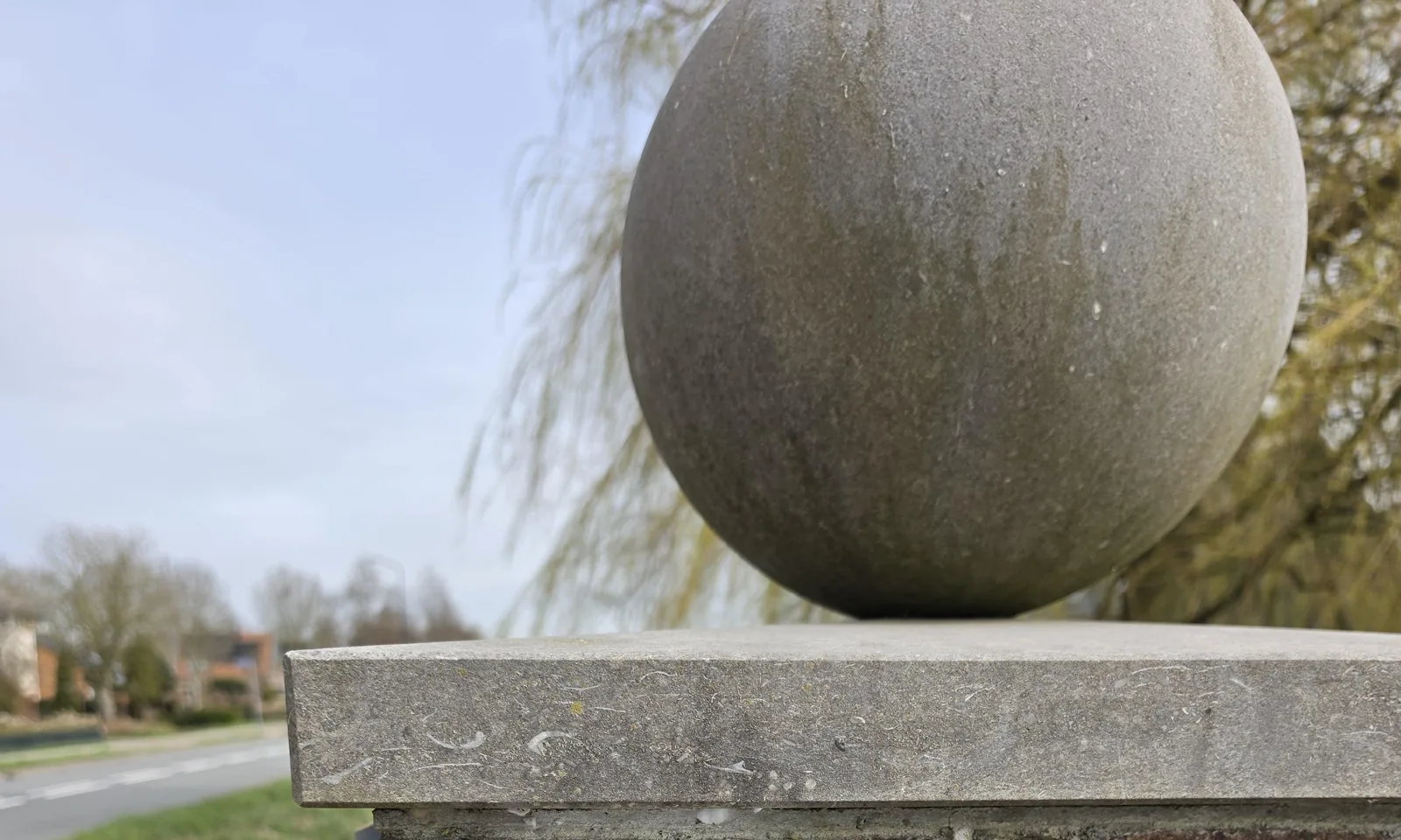 Bovenkant van de bakstenen poortpilaar met een hardstenen afdekplaat en een grote stenen bol, met een treurwilg, een sloot en de weg ernaast