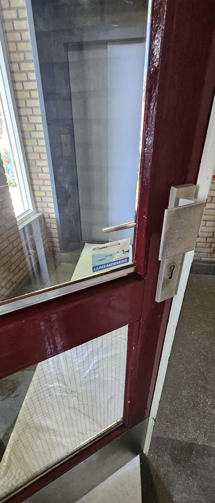 Donkerrode portiekdeur met glas en daarachter een sticker van CameraInstallatie.nl met de tekst camerabewaking 24 uur 7 dagen