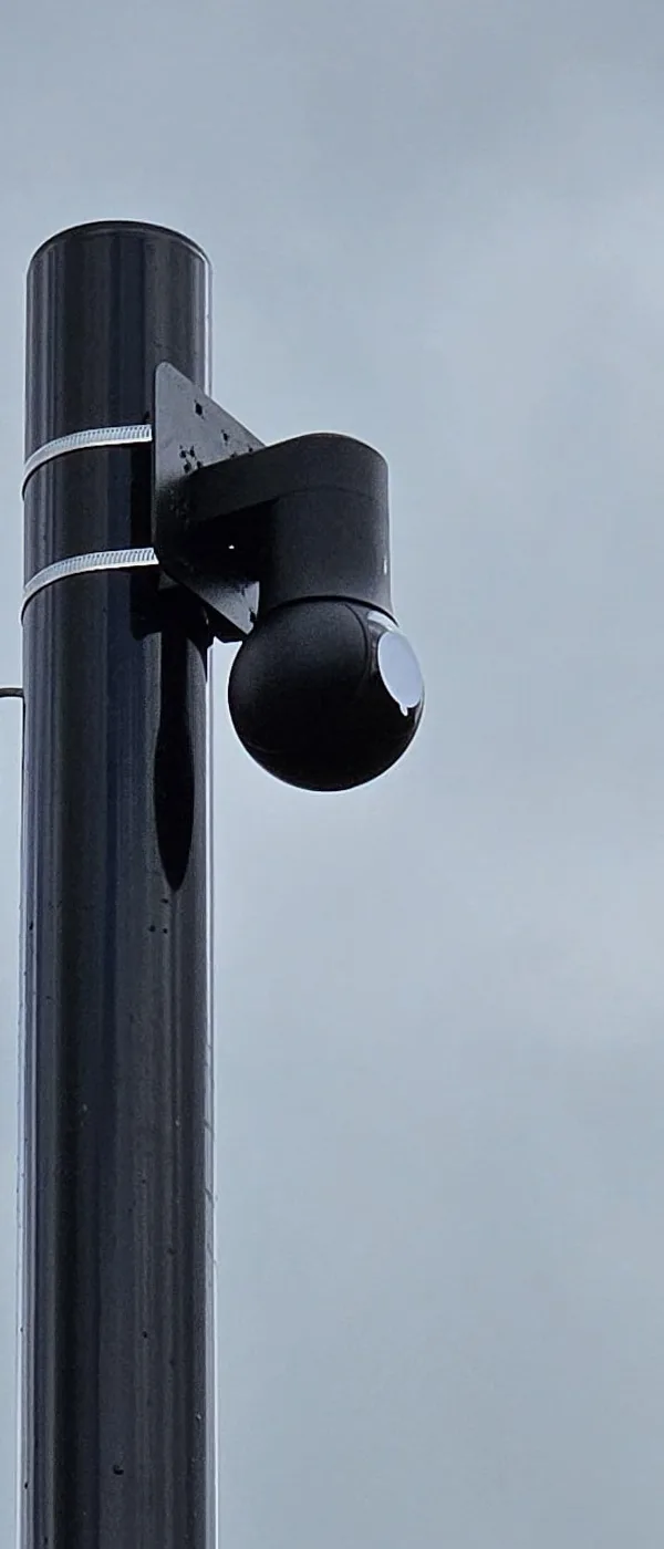 Zwarte PTZ-camera aan een beugel bovenaan een zwarte camerapaal, tegen een grijze lucht