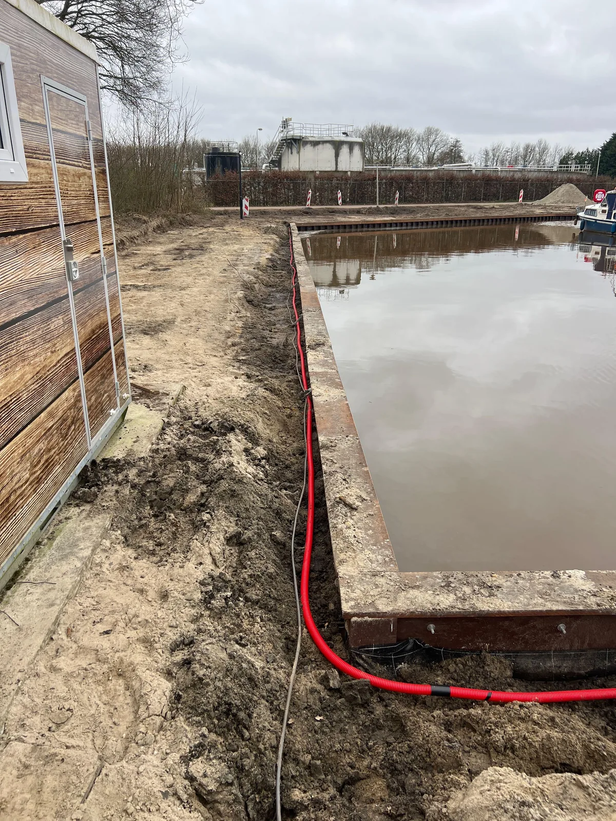 Open sleuf met een rode mantelbuis voor de glasvezel langs een houten gebouw en het water in Blokzijl