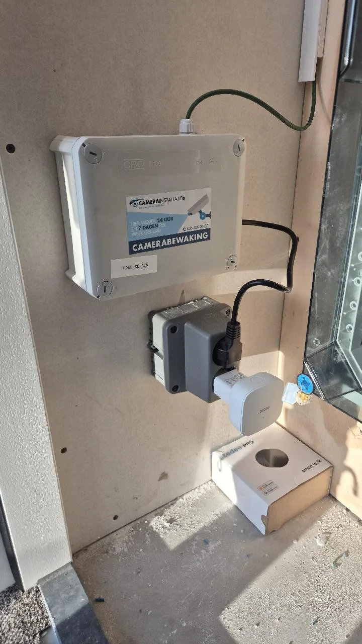Grijze aansluitdoos met de sticker van CameraInstallatie.nl en het label Tedee relais, met daaronder de Tedee-bridge en een doos Tedee PRO