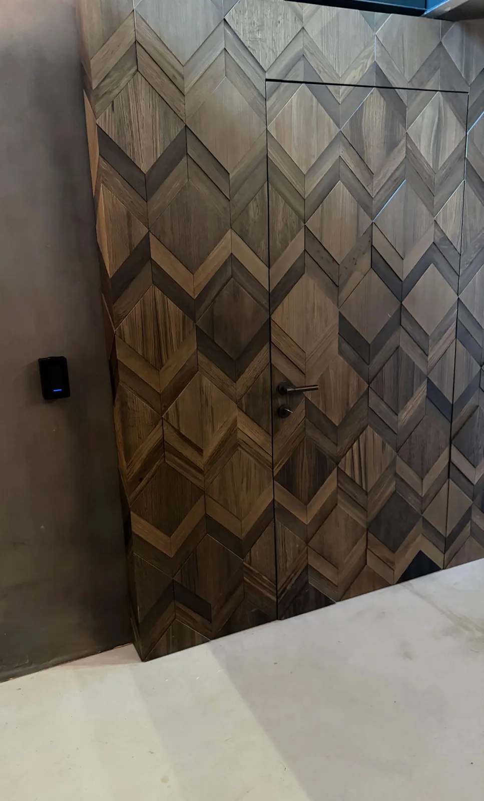 Zwarte UniFi-toegangslezer op een betonlook-wand, naast een deur die bijna onzichtbaar in een wand van houten ruitpanelen zit