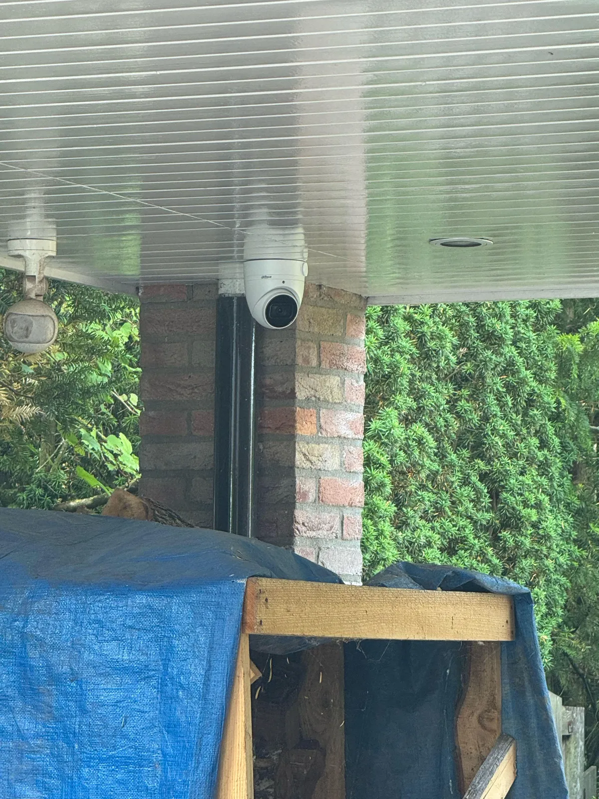 Witte turretcamera onder een overkapping bij een bakstenen pilaar, met groene haag en een blauw zeil op de voorgrond