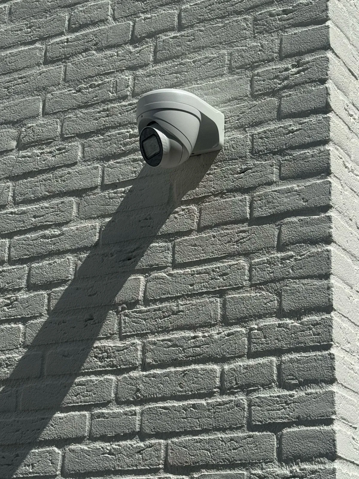 Witte turretcamera op een muurbeugel op de hoek van een lichte bakstenen gevel, met een scherpe schaduw
