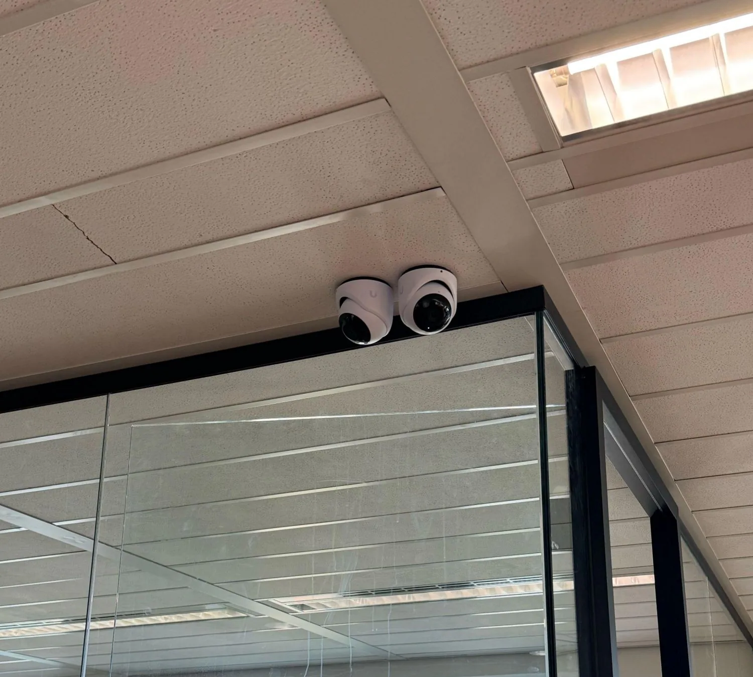 Twee witte UniFi-turretcamera's bovenop een glazen unit onder het systeemplafond van een kantoor