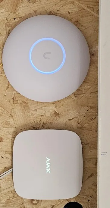 Wit UniFi-accesspoint en een witte Ajax Hub naast elkaar op OSB, bovenin de meterkast