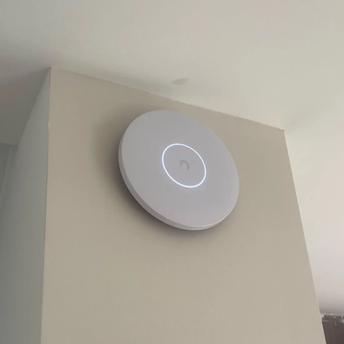 Rond wit UniFi-accesspoint met blauwe ring hoog op een lichte wand, vlak onder het plafond