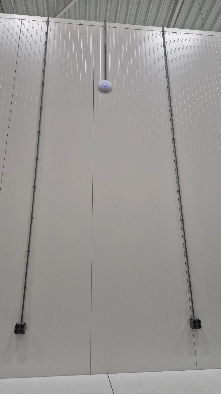 UniFi-access point midden op een hoge paneelwand, met links en rechts een kabelbuis naar beneden