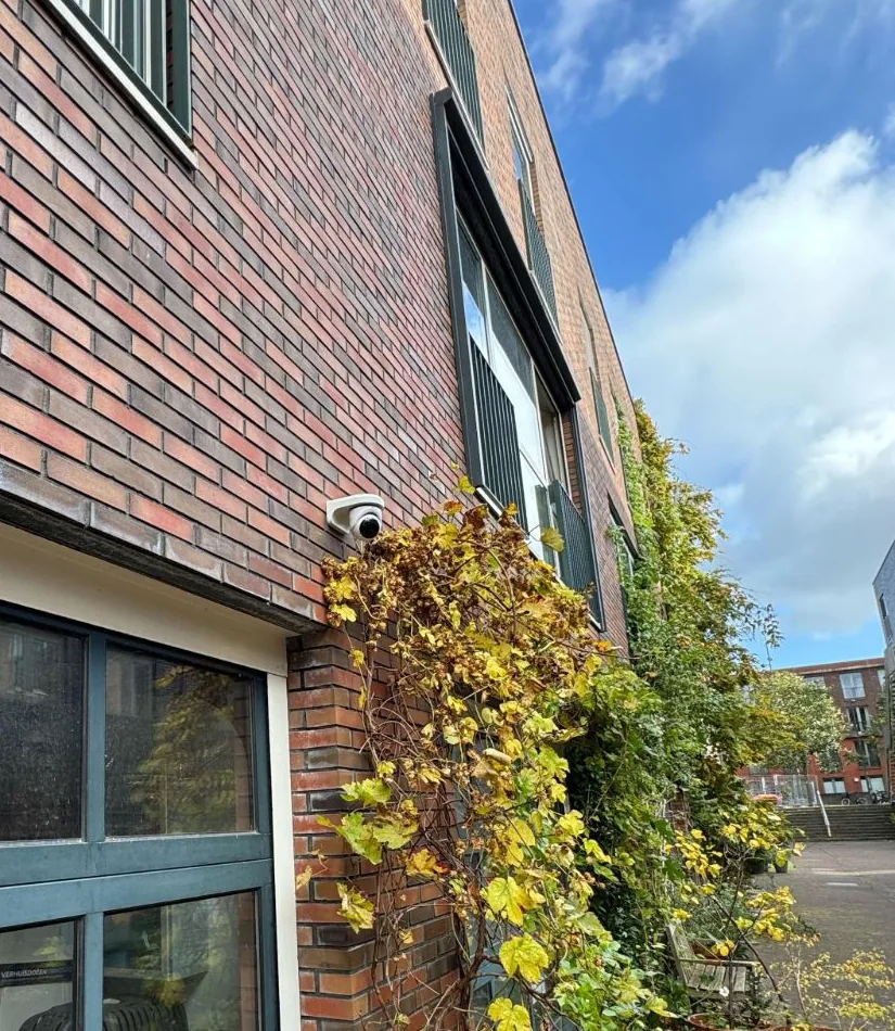 UniFi-domecamera op een bakstenen gevel, boven een glazen garagedeur en naast klimop