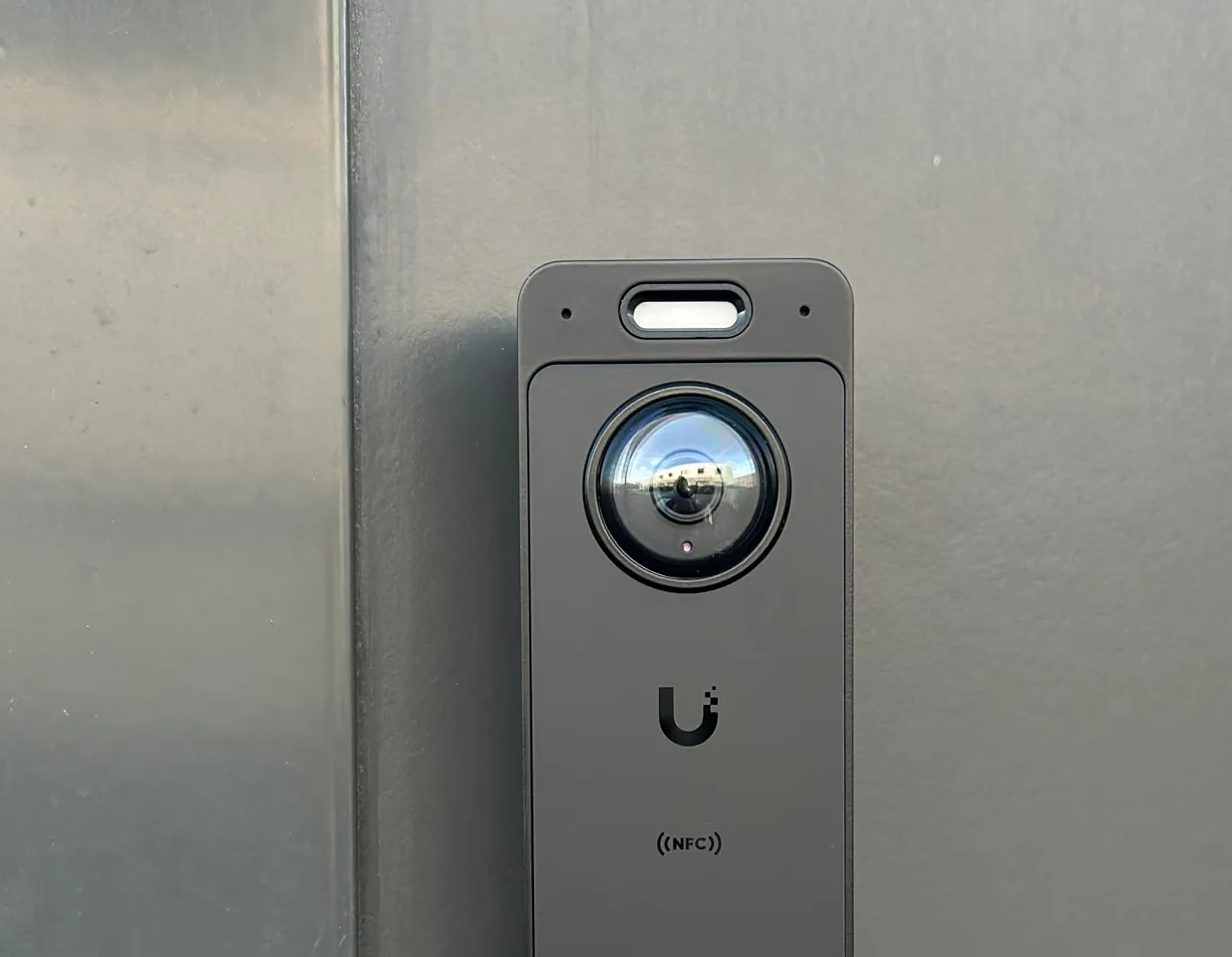 Zwarte UniFi-deurbel met camera, NFC-lezer en beldrukker met blauwe ring, recht op een donkergrijs aluminium kozijn