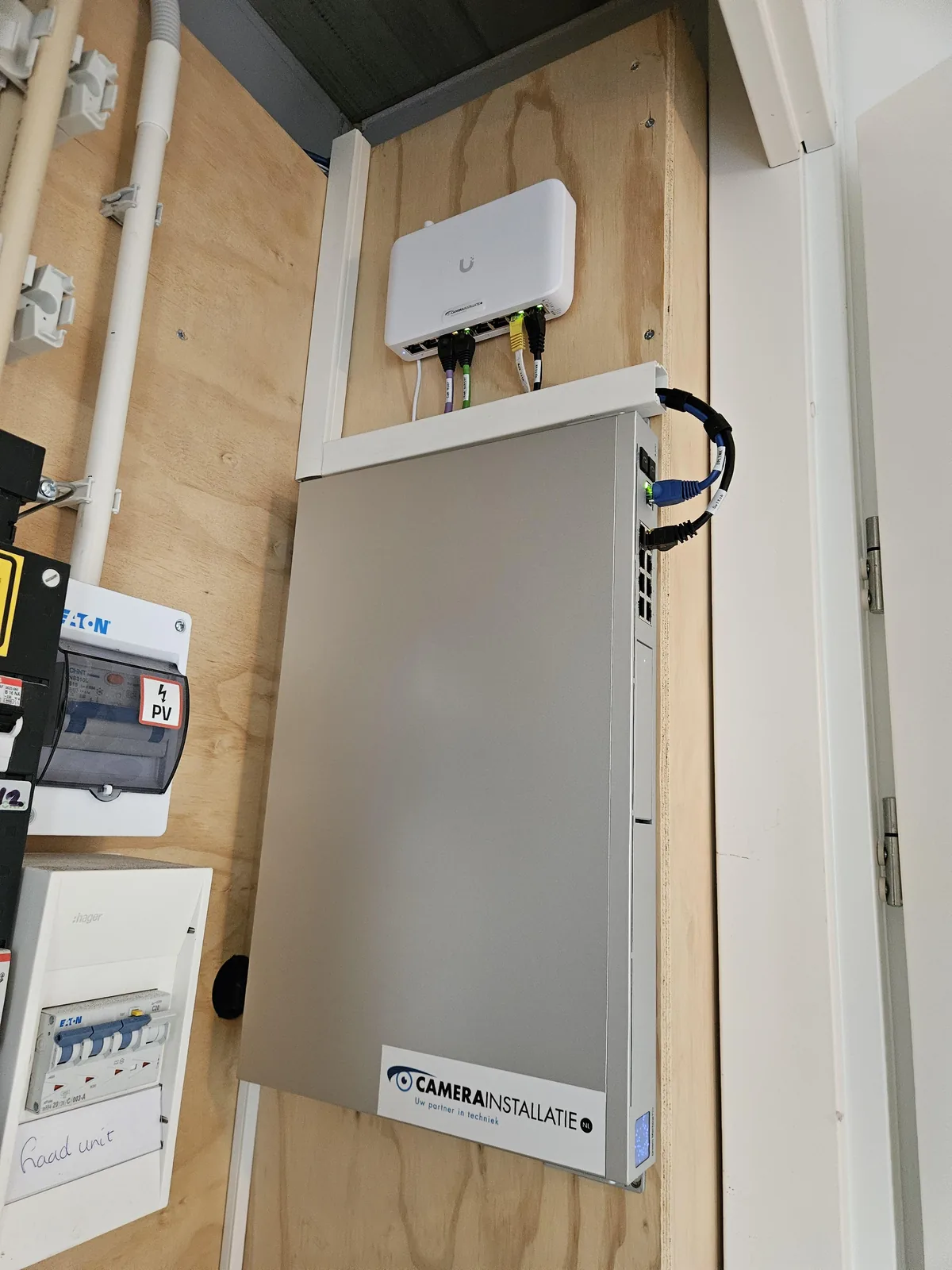 Grijze UniFi Dream Machine rechtop tegen de plaat in de meterkast van een woning in Amstelveen, met erboven een witte UniFi PoE-switch