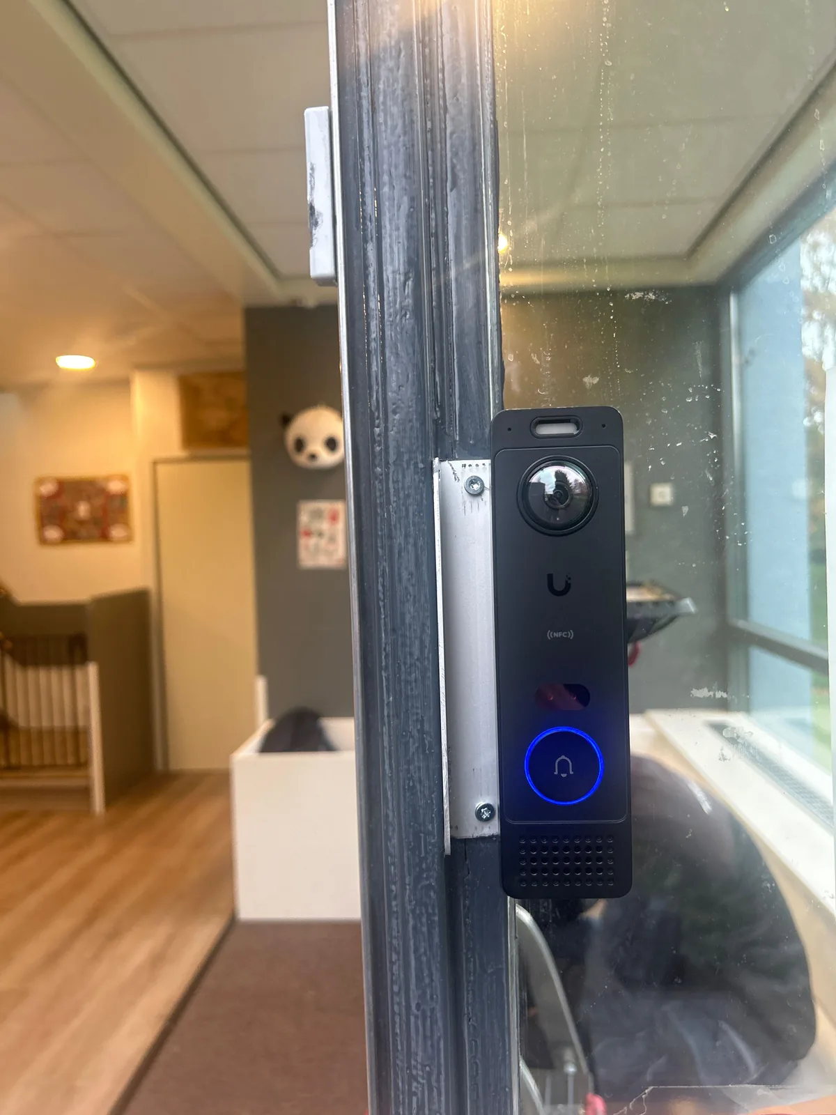 UniFi G6 Entry, een deurbel met camera en NFC-lezer, op een aluminium plaat tegen het kozijn van de gele deur van een kinderdagverblijf