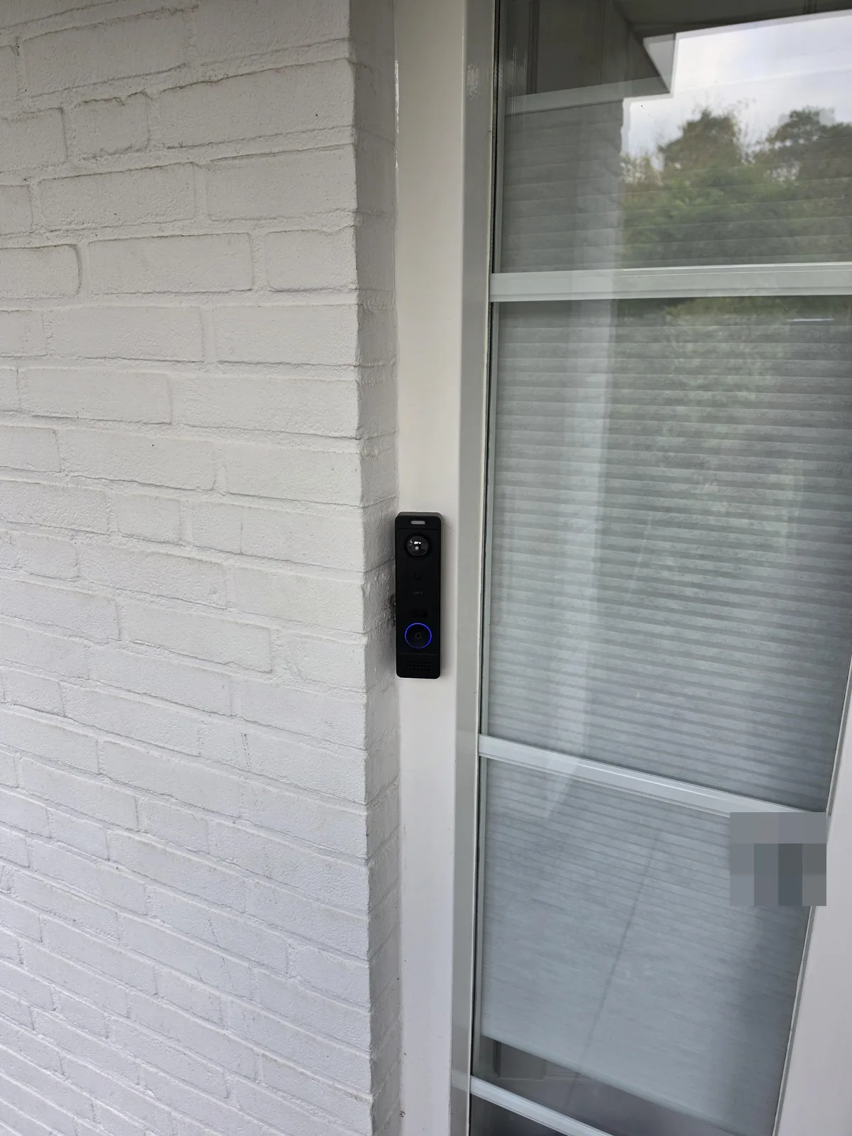 Zwarte UniFi G6 Entry-videodeurbel op het witte kozijn naast de voordeur, tegen witgeschilderde baksteen