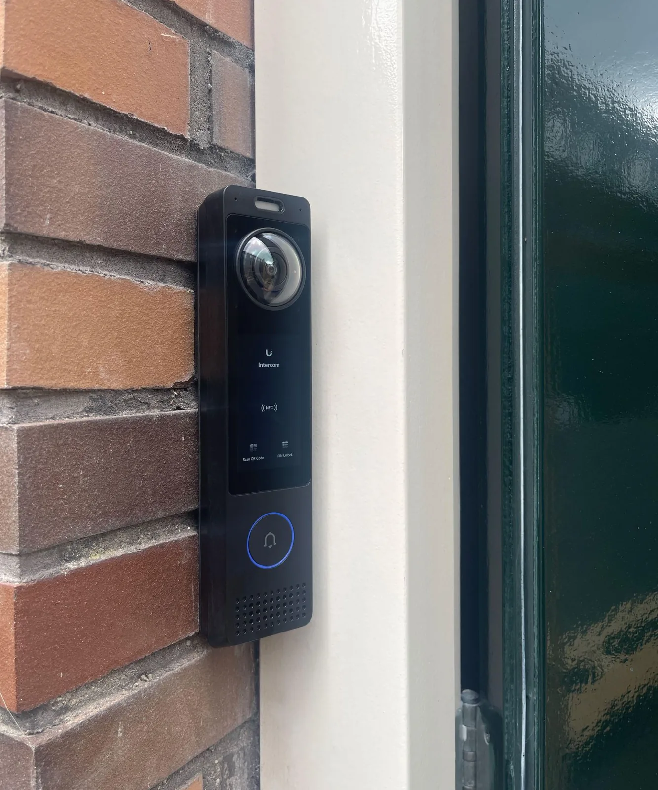 Zwarte UniFi G6 Pro Entry, een deurbel met camera en toetsfuncties op een bakstenen gevel naast het kozijn van de voordeur