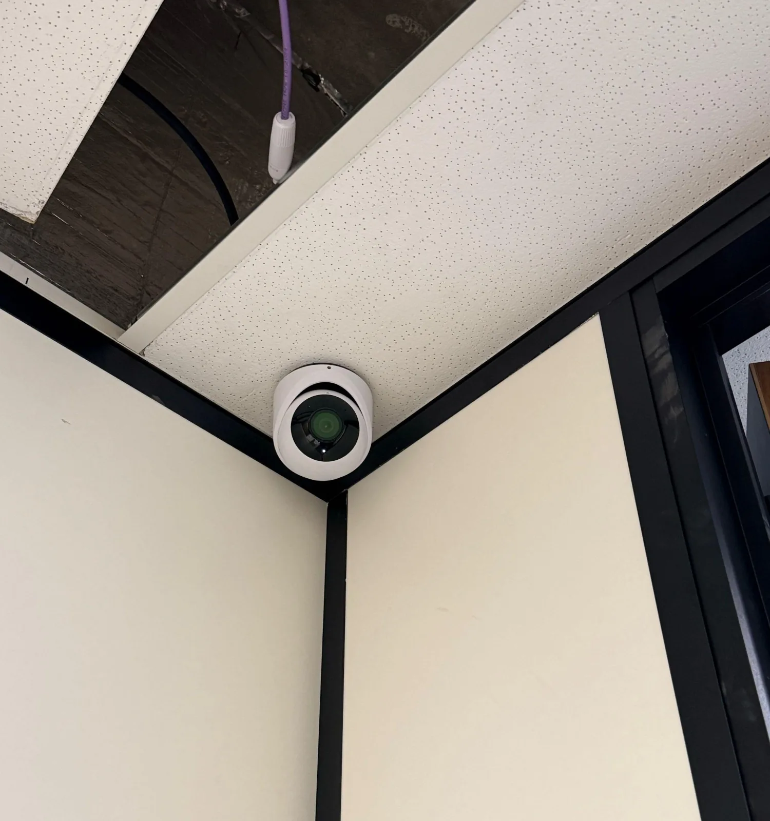 Witte UniFi-turretcamera van dichtbij in een hoek, met een paarse kabel uit het open plafond