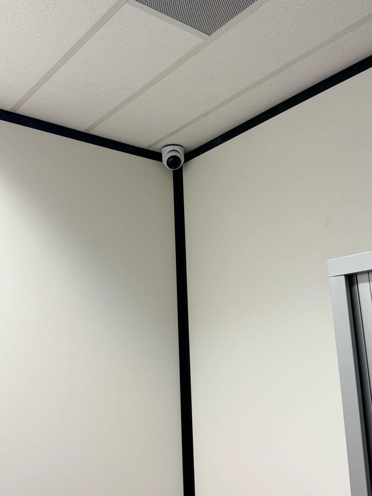 Witte UniFi-turretcamera in de hoek onder het systeemplafond, tussen zwarte profielen