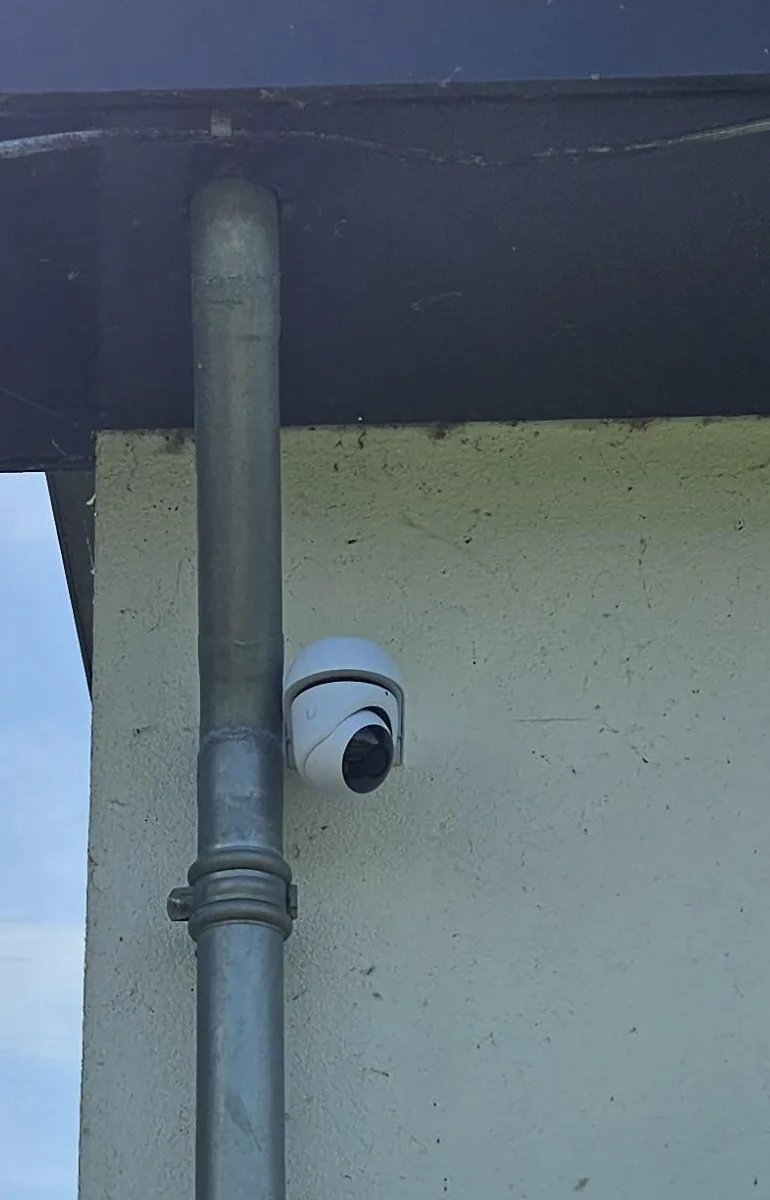 Witte UniFi G6 Turret-camera op een witte gevel onder een donkere dakrand, naast een regenpijp