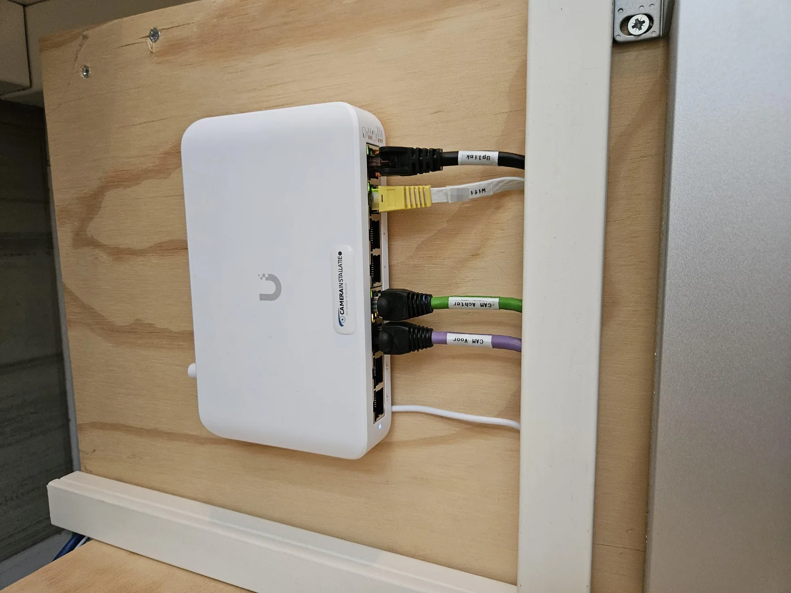 Witte UniFi PoE-switch met gelabelde kabels: Uplink, wifi, CAM achter en CAM voor