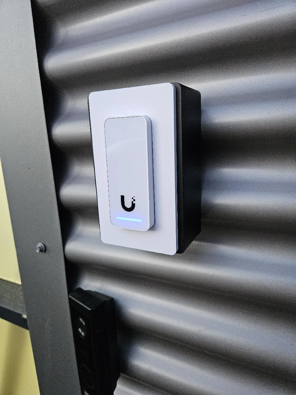 UniFi Access Reader G2 van opzij, op de zwarte junction box tegen de golfplaat