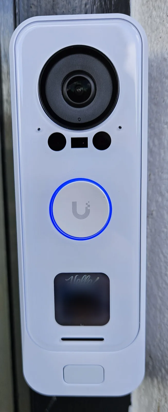 Witte UniFi-videodeurbel met blauwe ring om de belknop, op het zwarte kozijn van de voordeur