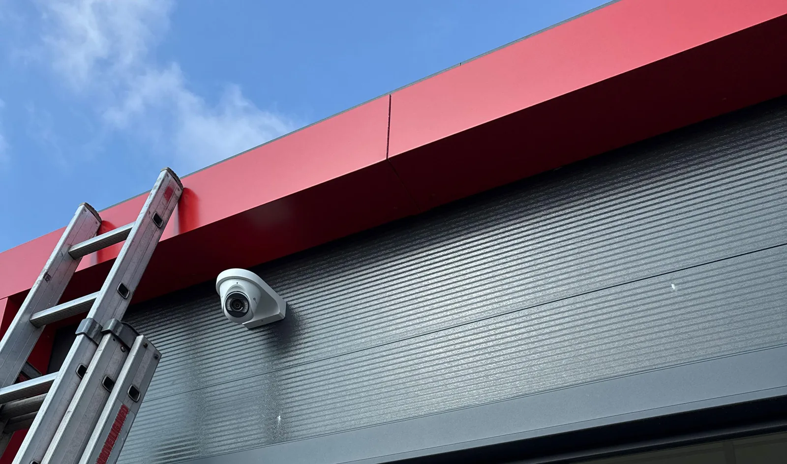 Witte VIGI-turretcamera op een hoekbeugel tegen een donkergrijze gevel, vlak onder een rode dakrand, met links een aluminium ladder tegen de gevel