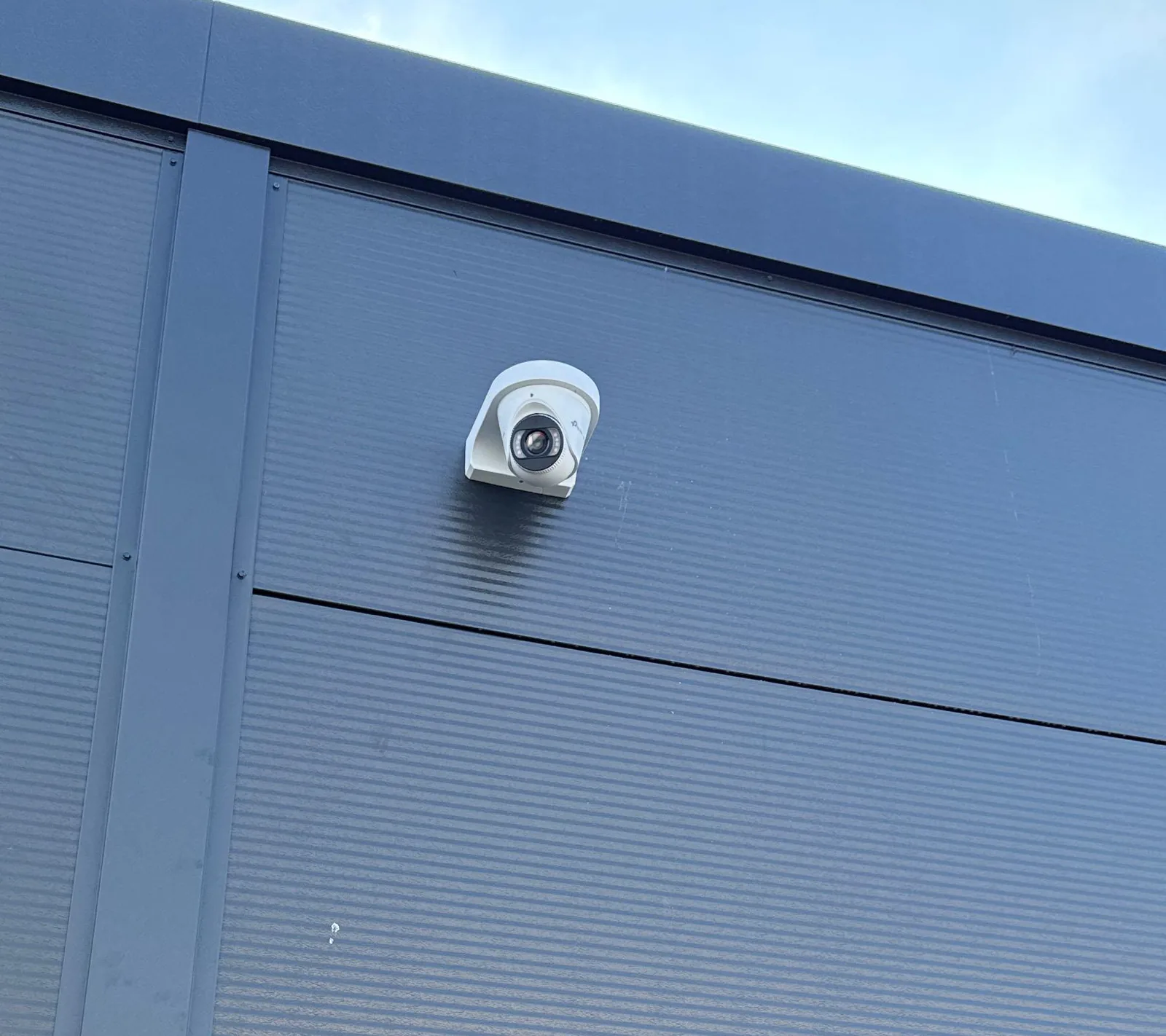 Witte VIGI-turretcamera op een beugel hoog op een donkergrijze gevel van sandwichpanelen, onder de dakrand