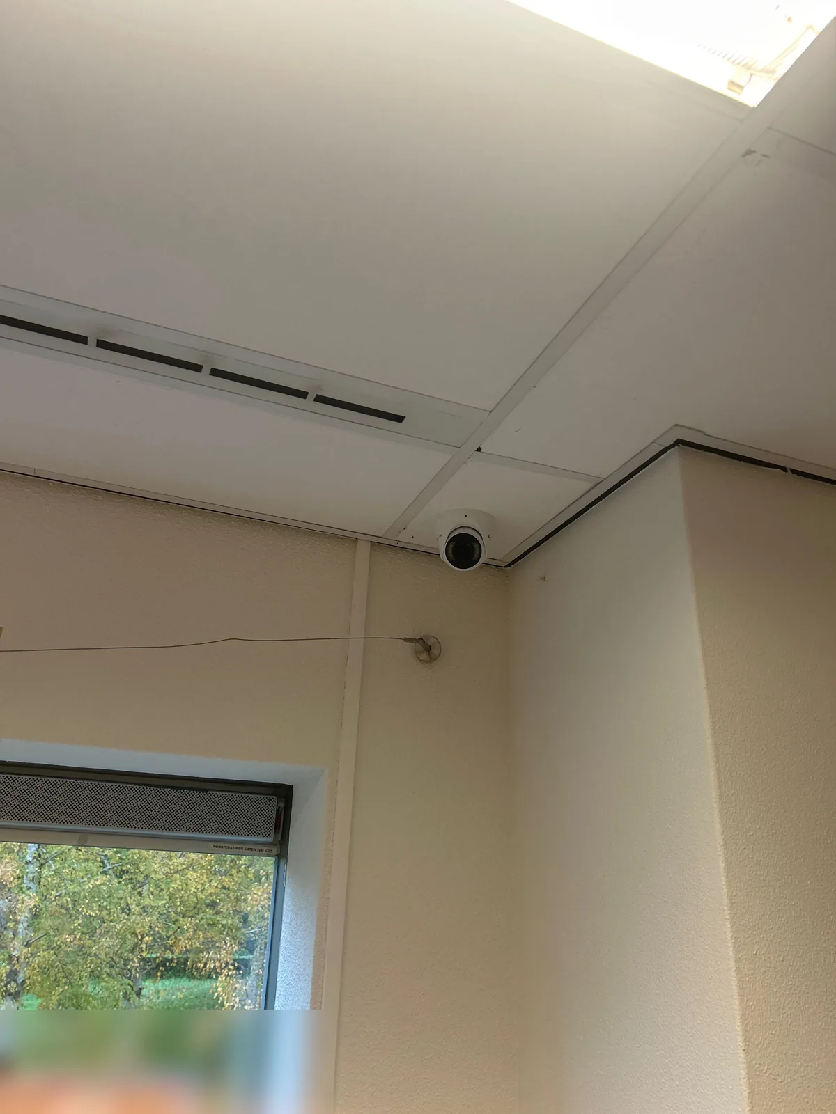 Witte VIGI-camera hoog in de hoek van een groepsruimte, tegen het systeemplafond naast het raam