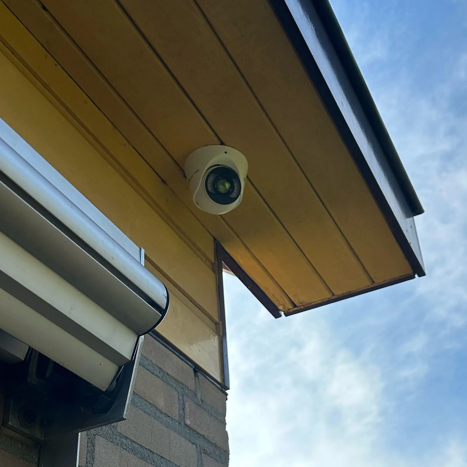 Witte VIGI-turretcamera onder de gele houten overstek, naast de kast van het zonnescherm, tegen een blauwe lucht