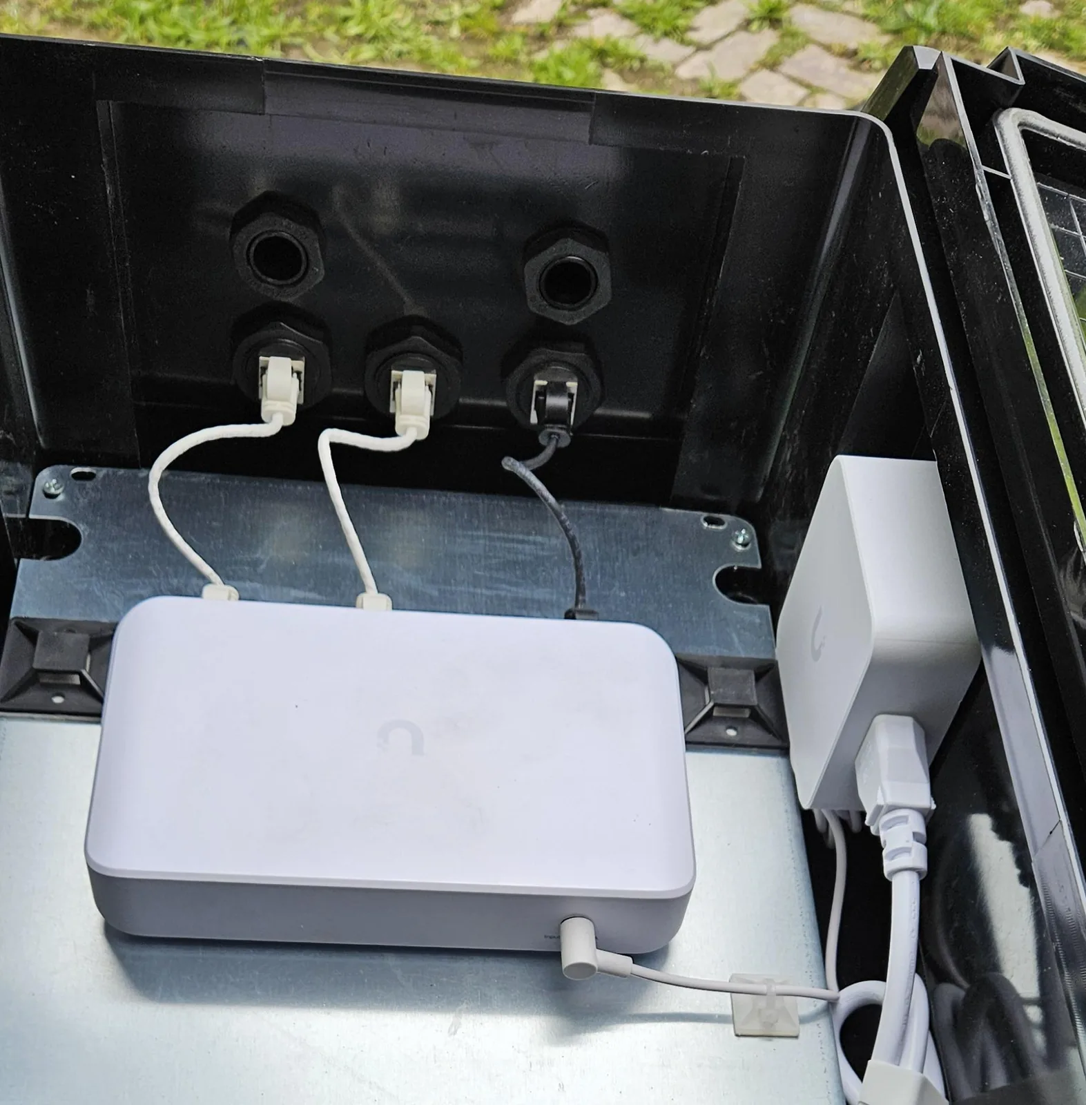 Netwerkkabels die via waterdichte doorvoeren in de wand van een kunststof kast binnenkomen en met korte patchkabels op een UniFi-switch zitten