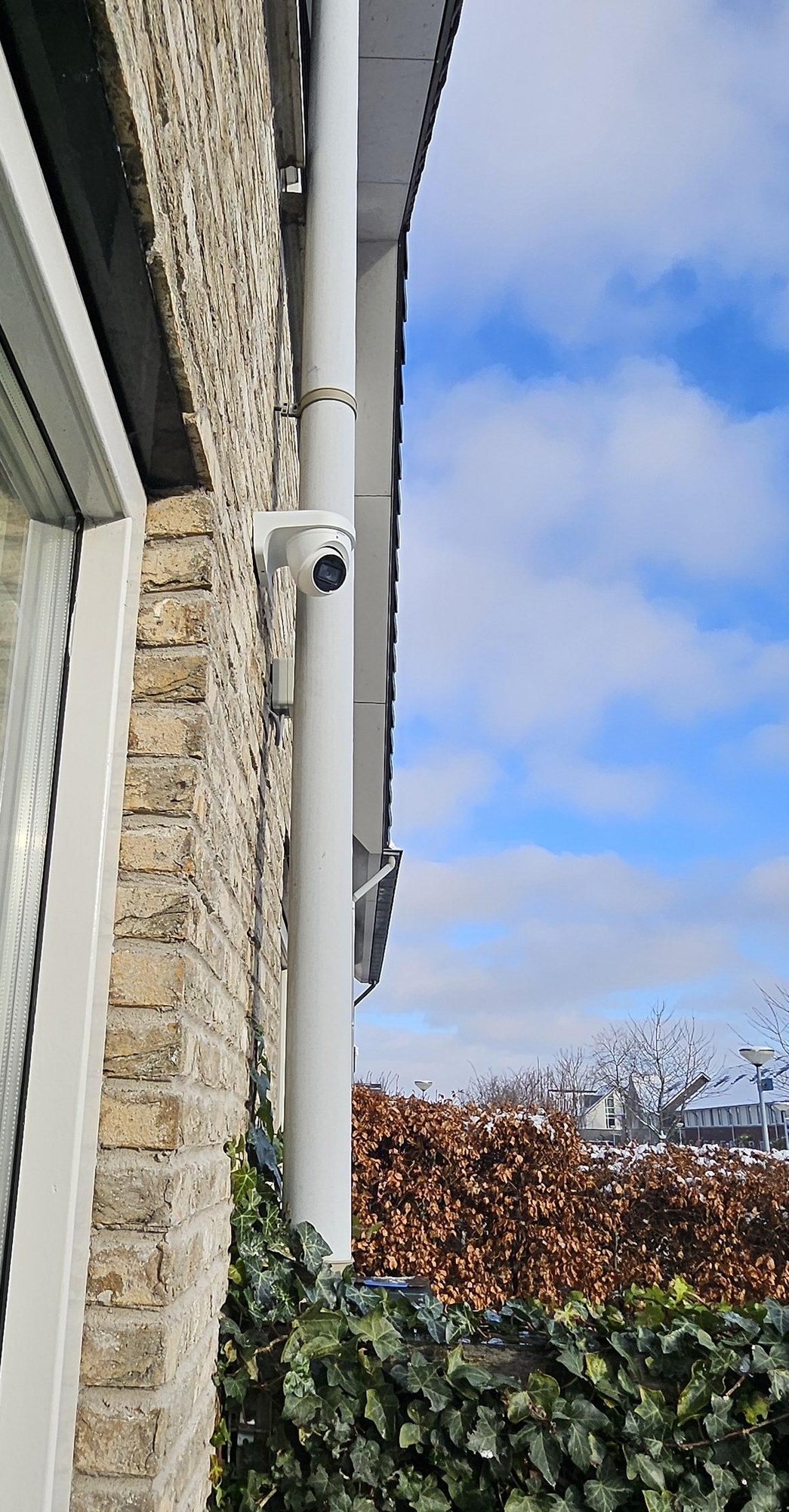 Witte camera hoog op de bakstenen gevel naast een witte regenpijp, met blauwe lucht