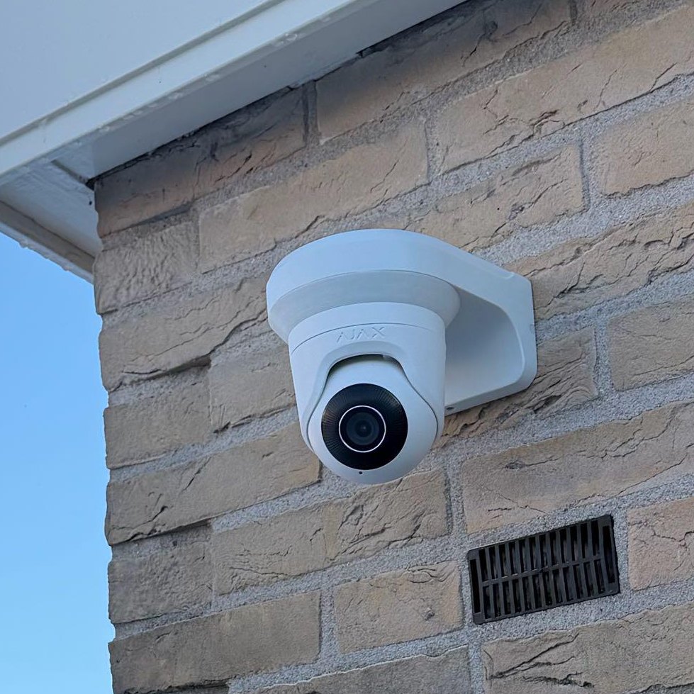 Witte camera op een witte muurbeugel tegen de bakstenen zijgevel, met een ventilatierooster ernaast