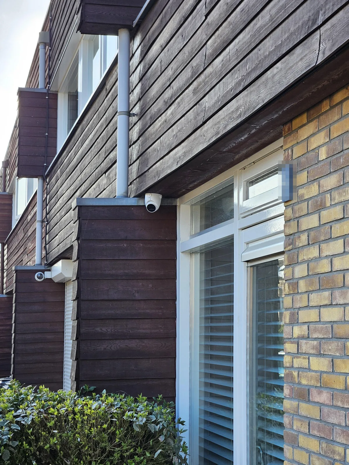 Witte Dahua-camera onder het houten overstek van een woning in Almere, op de erker naast het raam, zonder zichtbare kabel