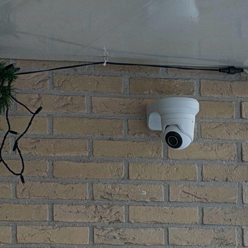 Witte turretcamera op een beugel tegen de bakstenen muur, met de kabel netjes onder de overkapping langs