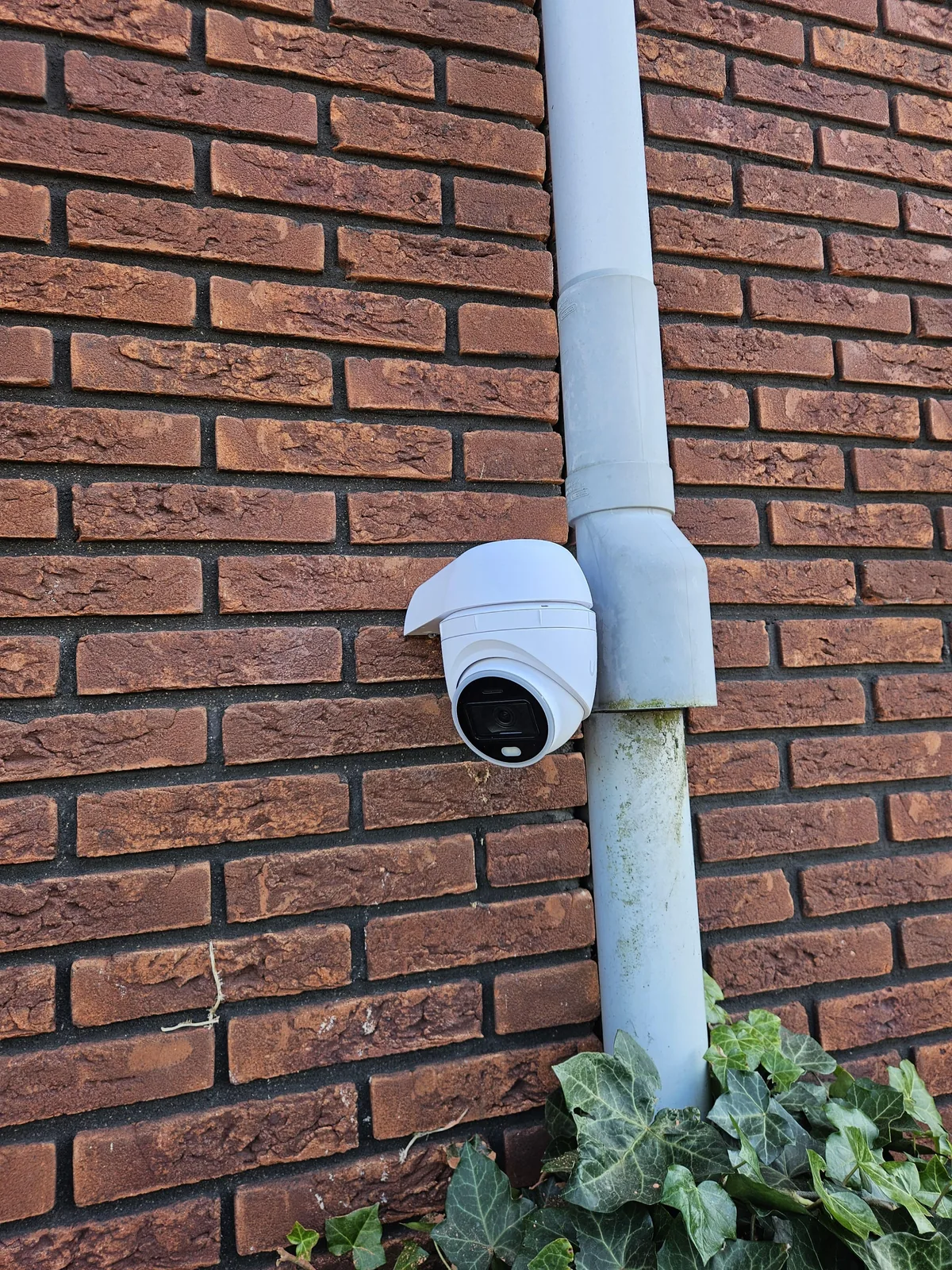 Witte UniFi AI Turret op een muurbeugel tegen de rode bakstenen gevel aan de kant van de achtertuin
