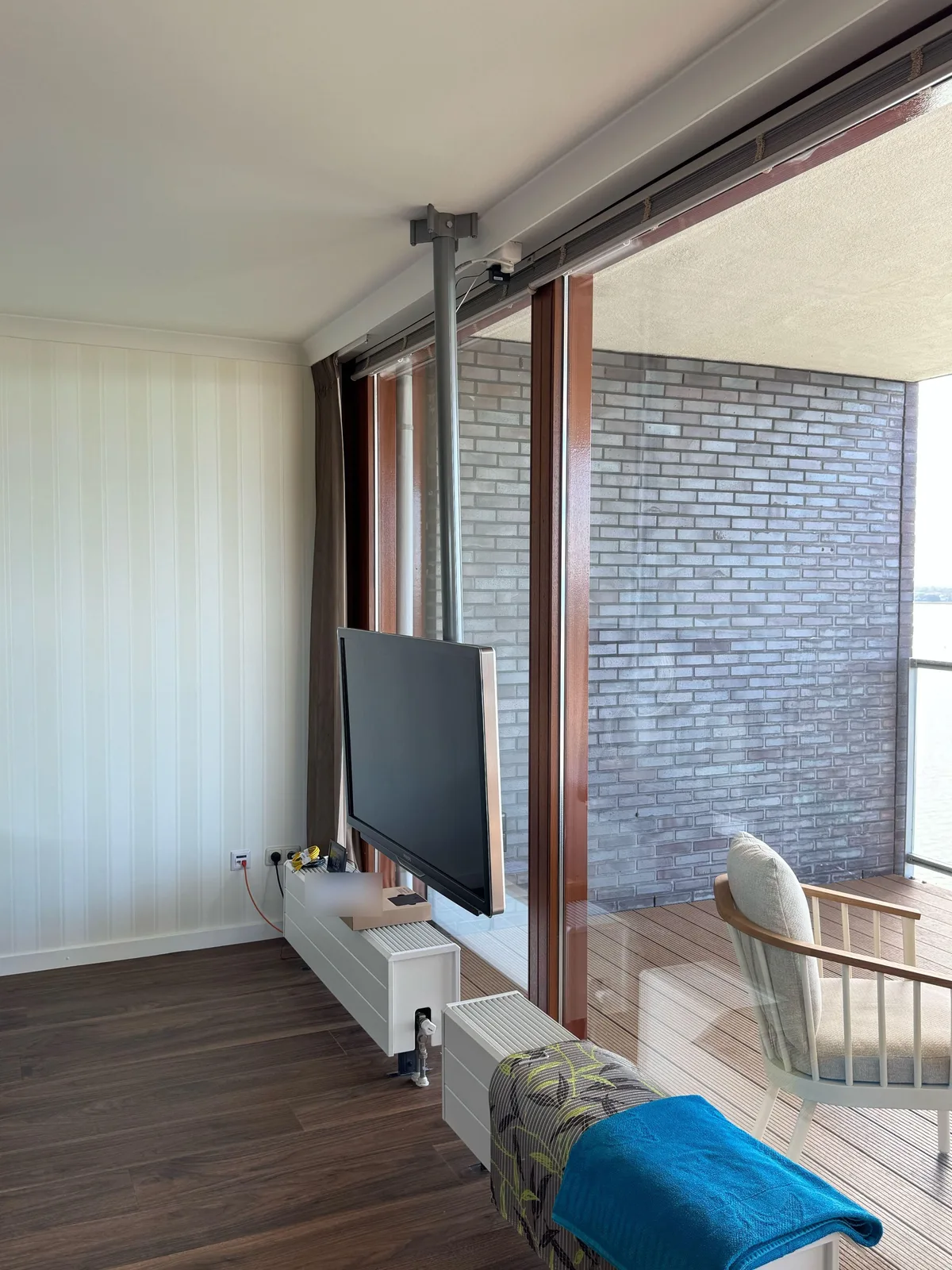 Lichte woonkamer met een tv aan een paal vanaf het plafond, naast een groot raam met balkon en uitzicht op het water