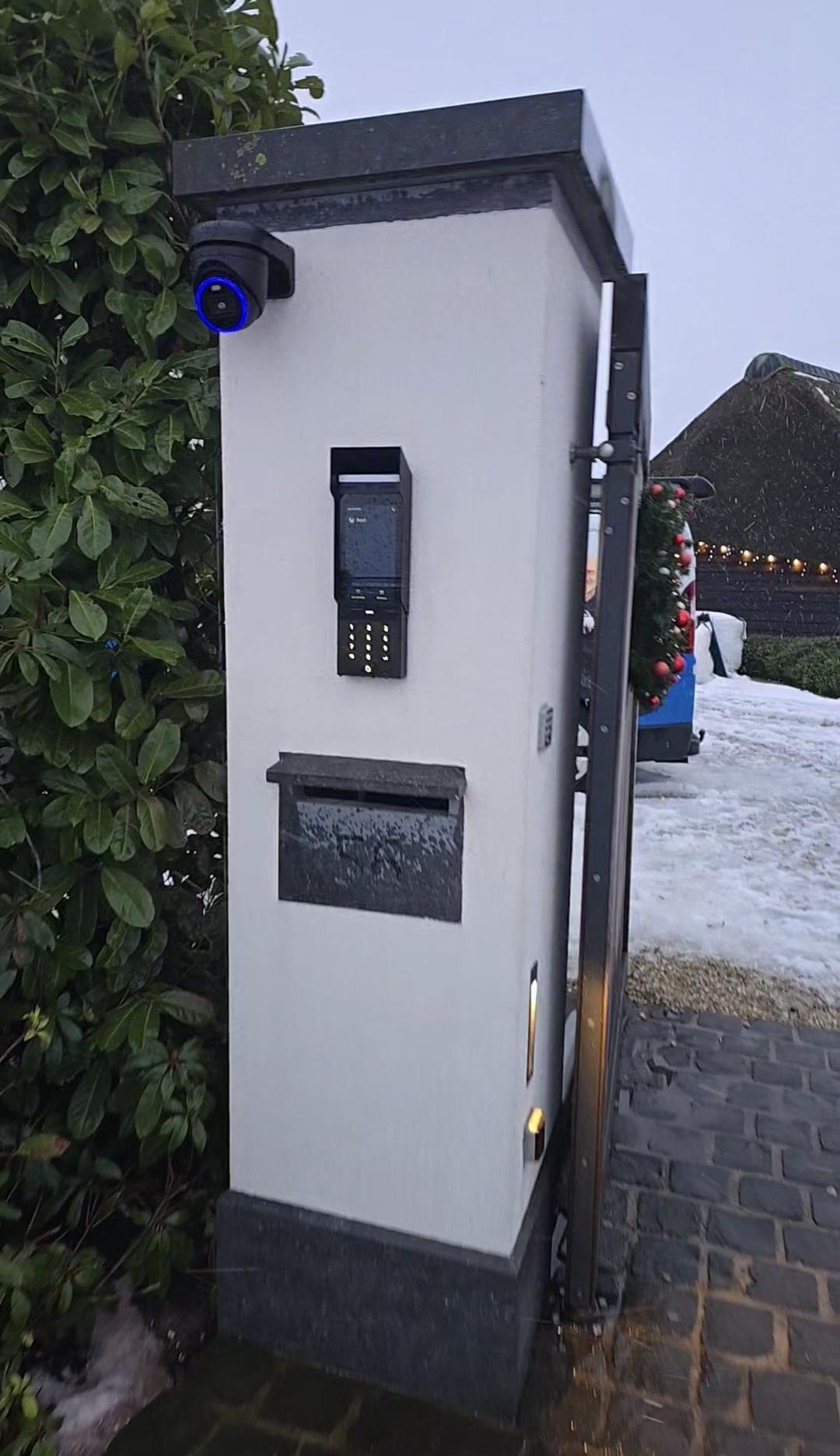 Witte zuil bij de oprit met bovenop een camera, daaronder de intercom en de brievenbus