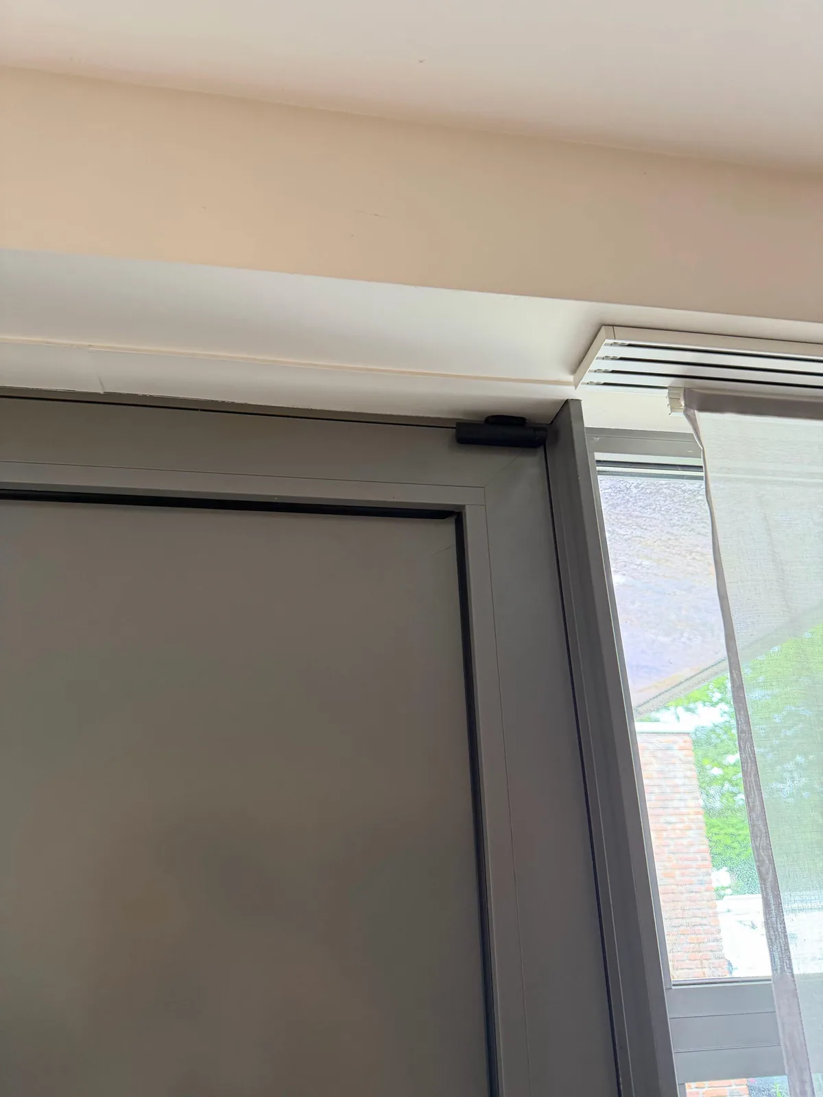Zwart Ajax-contact boven in een grijs deurkozijn, naast een raam met een vitrage, bij een woning in Kropswolde