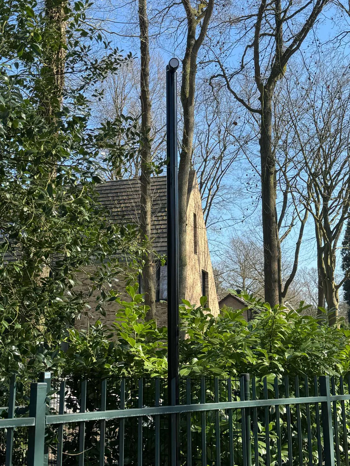 Slanke zwarte camerapaal achter het hek, tussen de bomen tegen een blauwe lucht